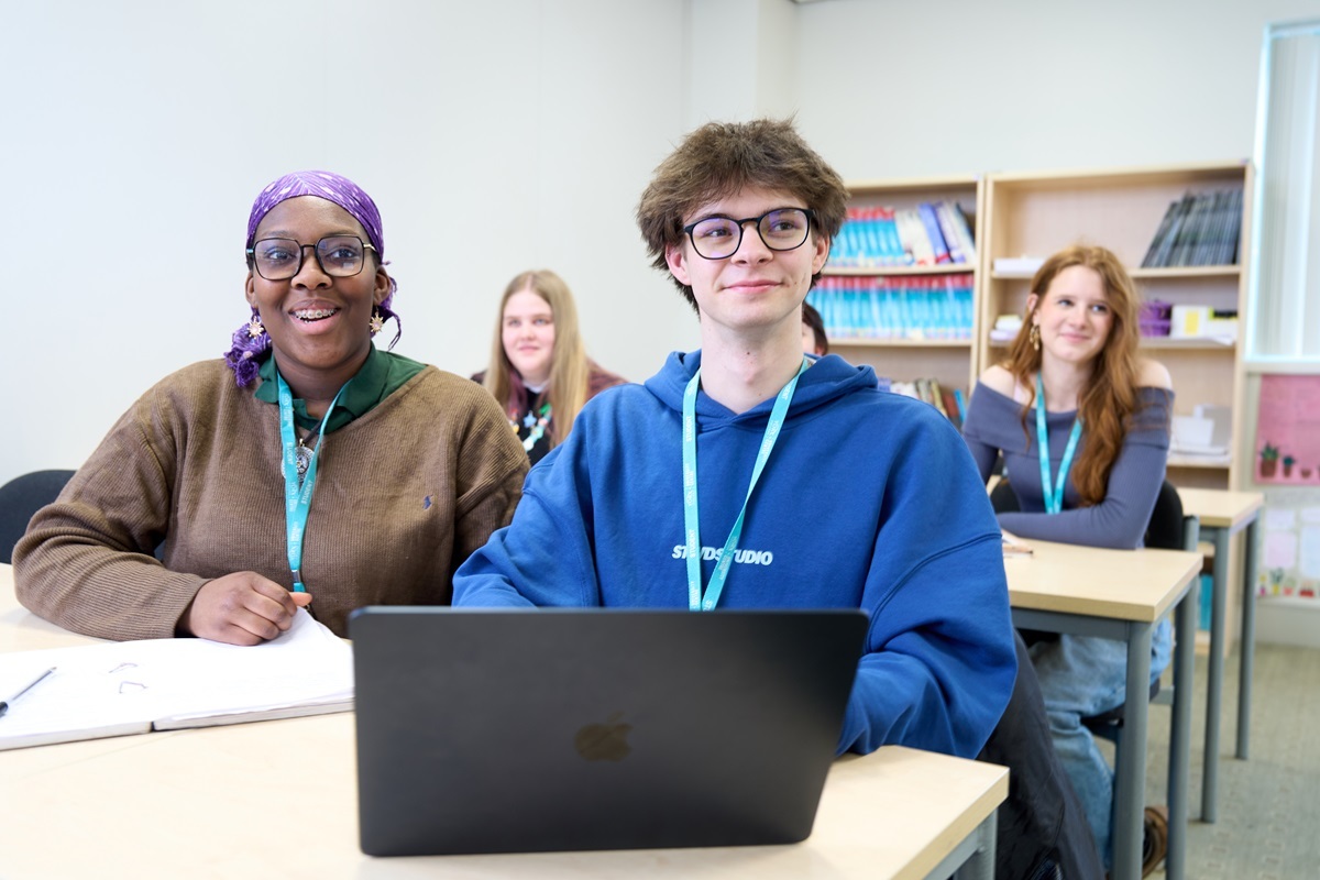 A Male student with a laptop and a female student with a notebook looking to the front of a classroom