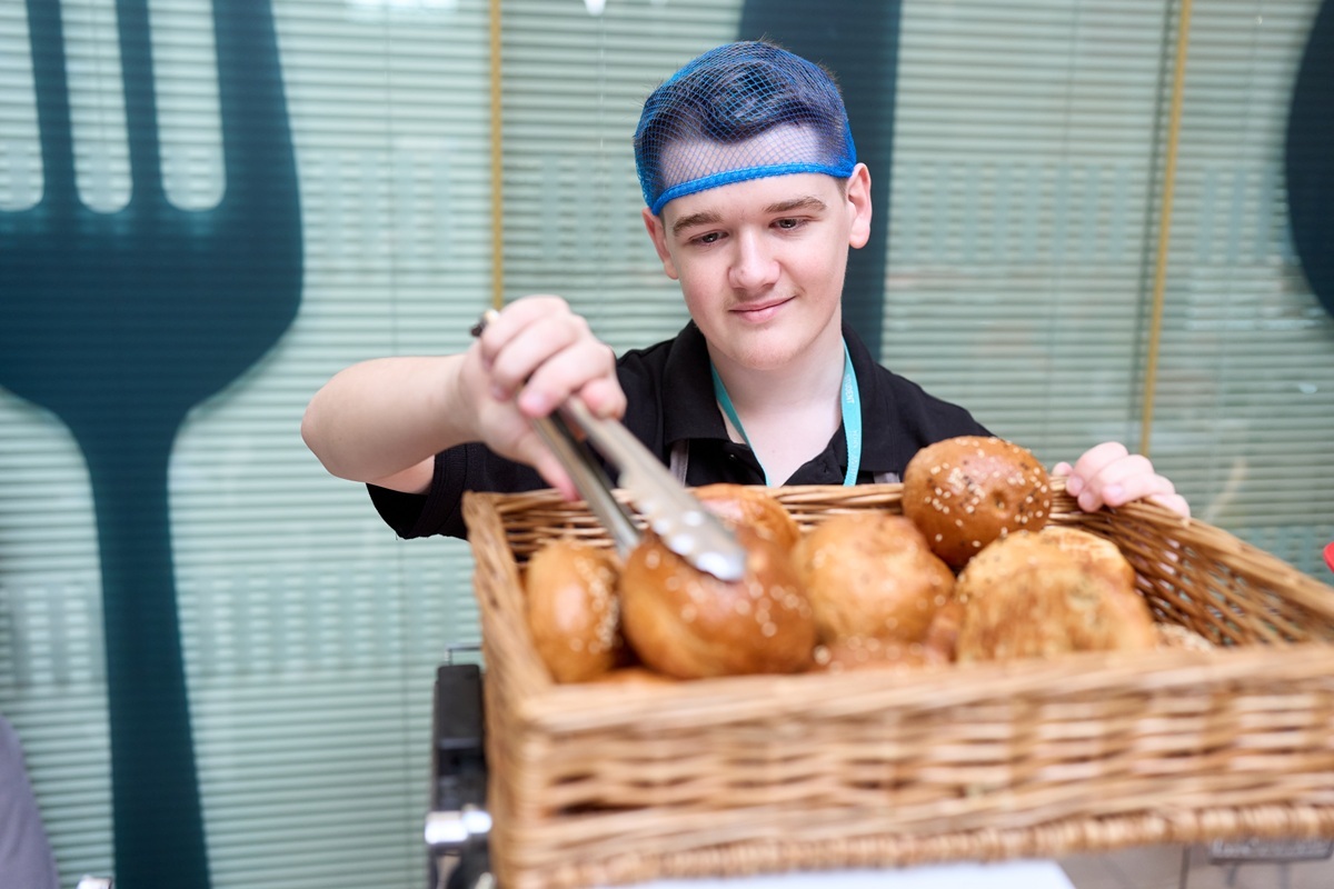 A hospitality student is using serving tongs to take a bread roll from a basket of seeded bread rolls