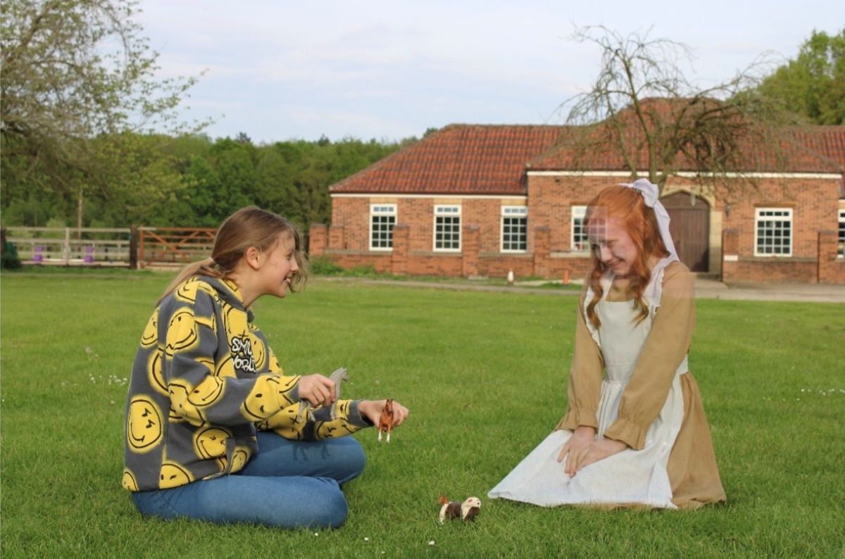 A modern teenage girl playing with toy horses in a big garden with a girl in old fashioned clothing whose head you can see part of the background through as if a ghost