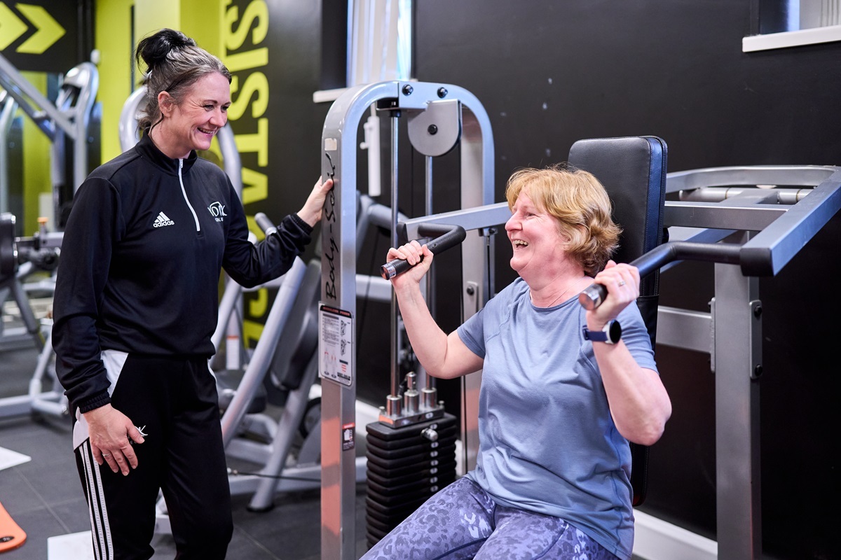 A woman performs a seated shoulder press and smiles at the tutor