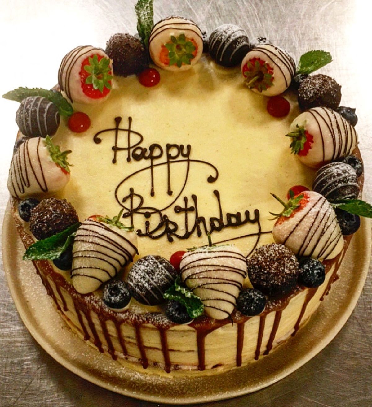 A yellow cake with Happy Birthday piped on top with icing and white chocolate covered strawberries truffles individual chocolates and blueberries around the circumference edge