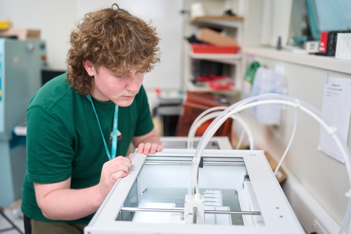An Engineeing student is looking into the top of a 3 D printer to check progress