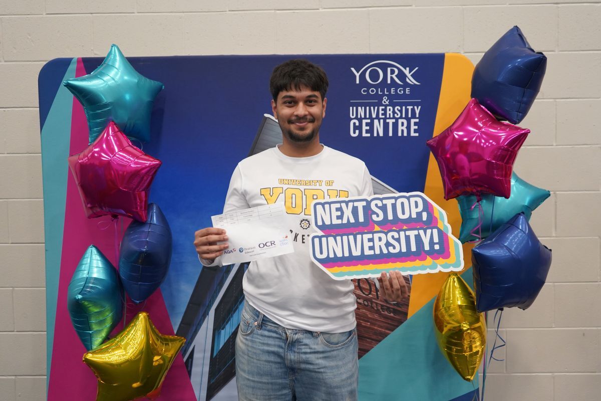 Azaan Iyer celebrates his A Level grades holding his results and a Next Stop University sign in front of banner with the York College building on it and surrounded by star shaped balloons