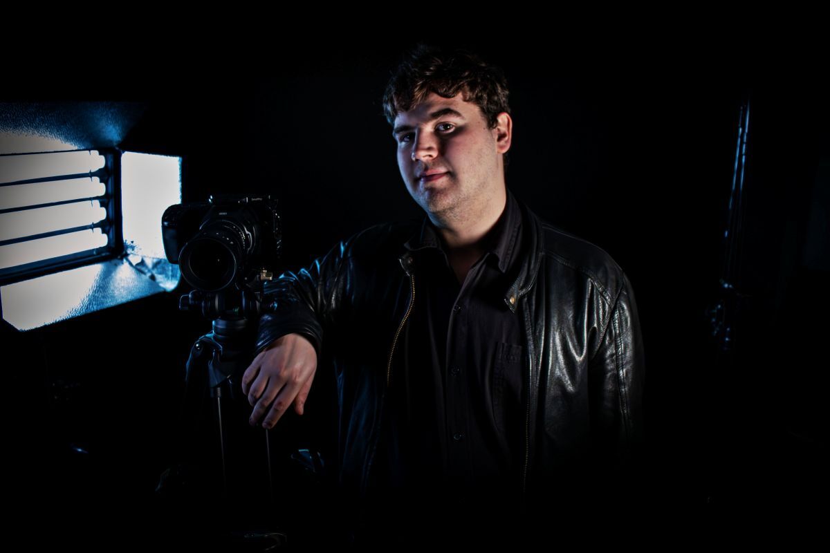 Dark artistic shot of Sam Matten leaning on a camera tripod with light from a vent illuminating his face
