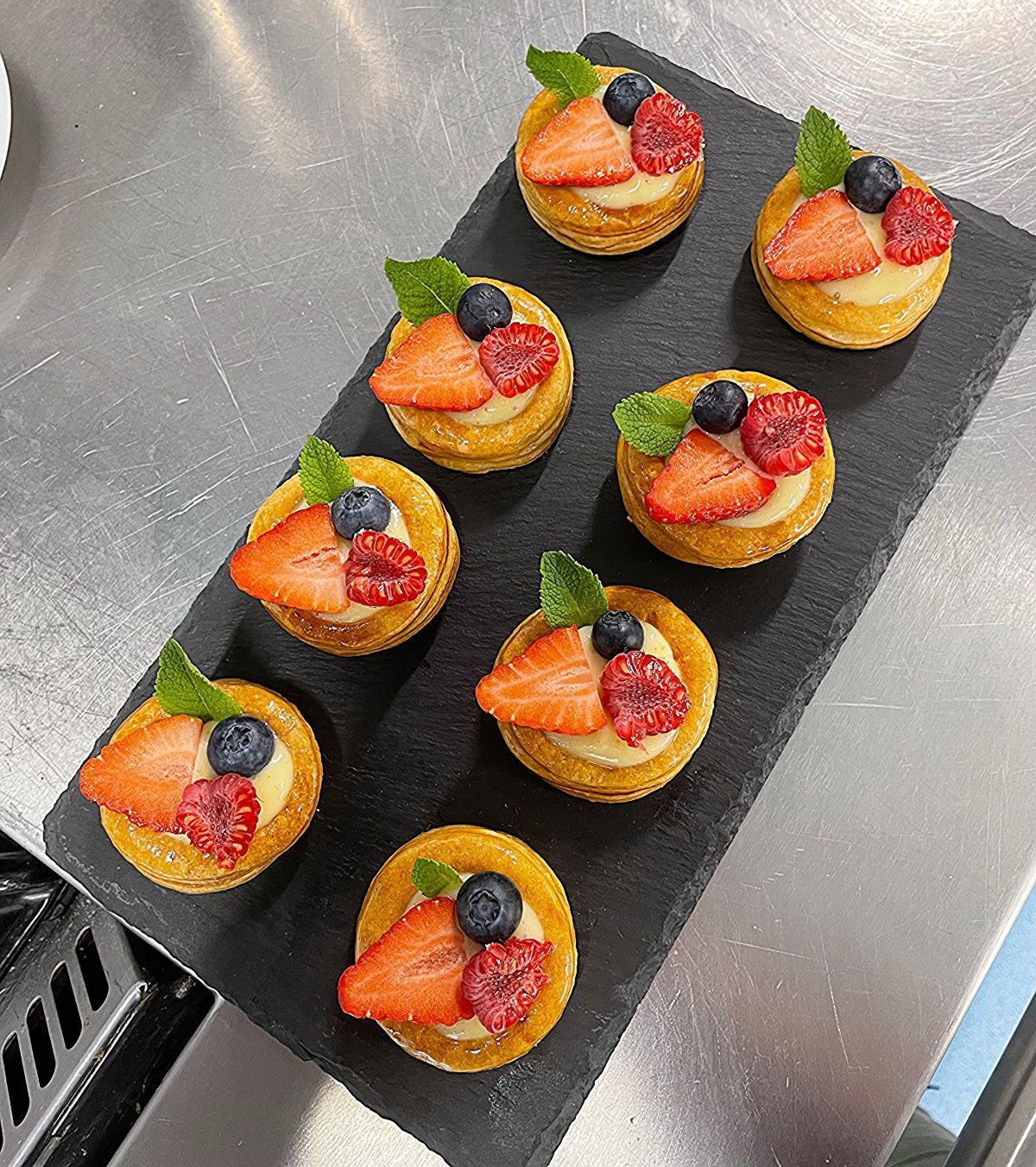 Eight identical round pastry desserts filled with mousse and half a strawberry a raspberry a mint leaf and a blueberry on top