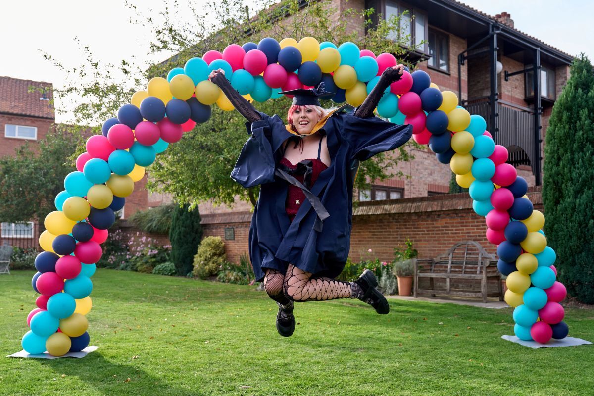 Female graduate jumps with both feet off the floor in front of the mutli coloured balloon arch in the front grounds at the Merchant Taylors Hall