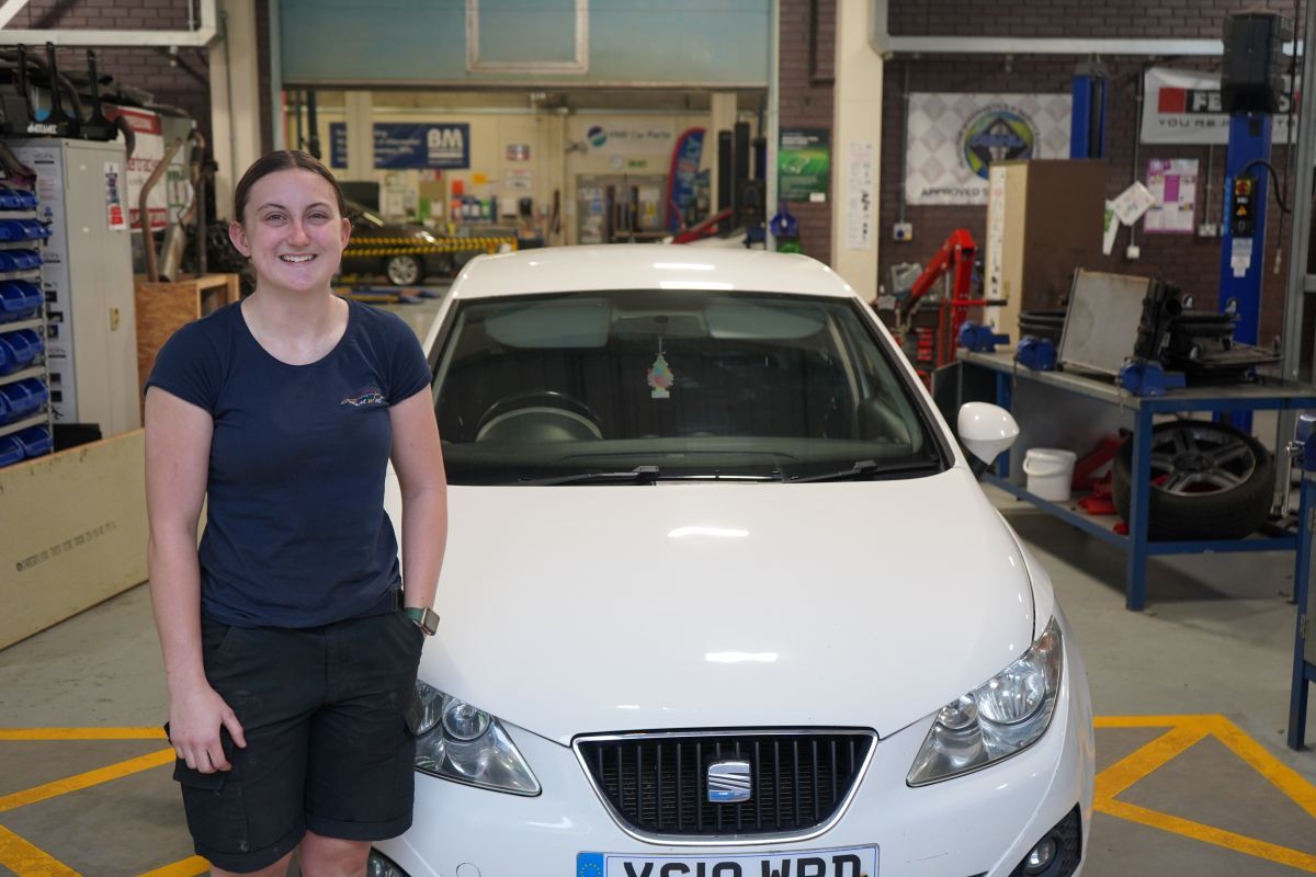 Georgina Richardson stands alongside a white Seat car in York Colleges Motor Vehicle workshop