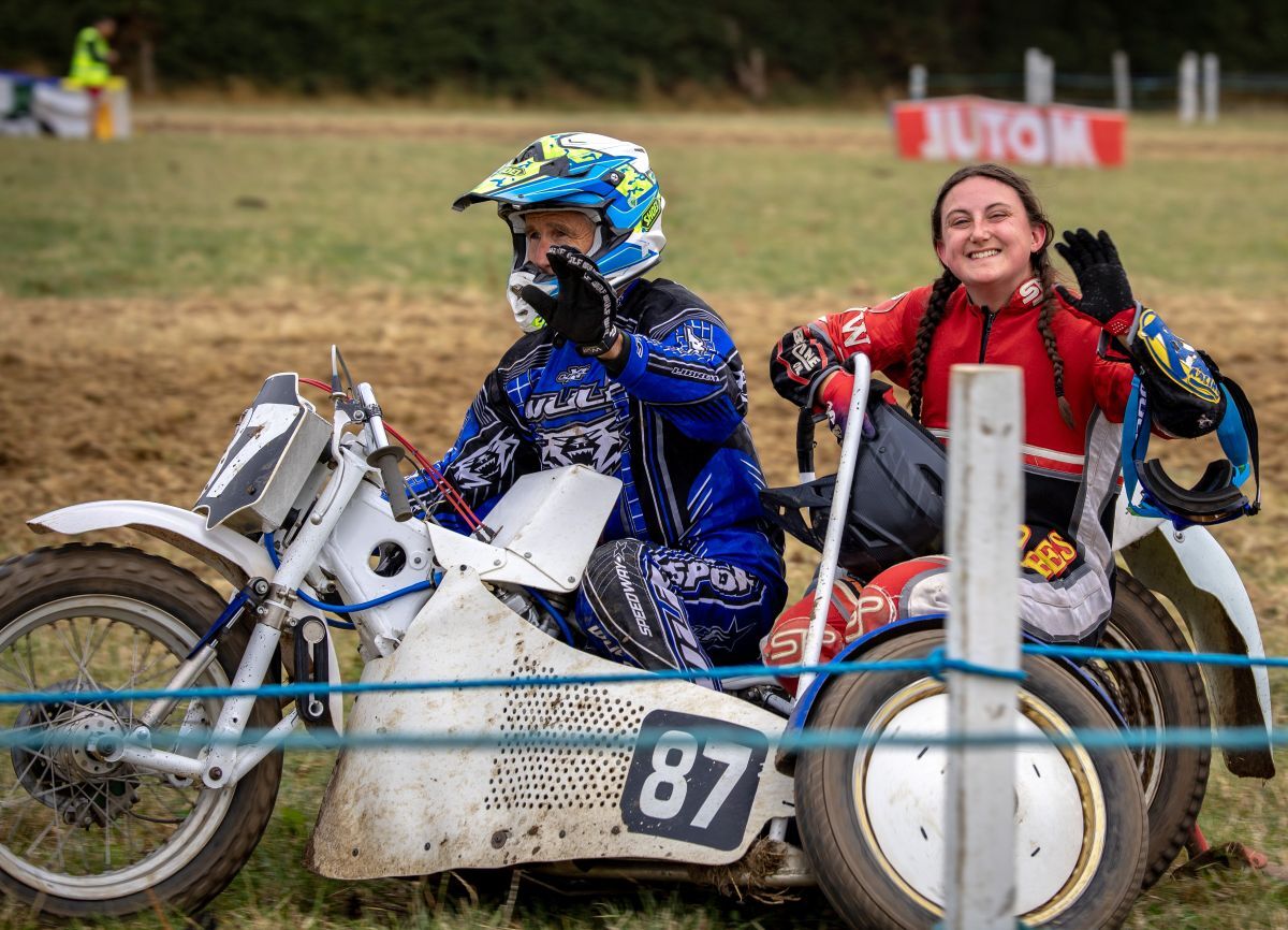 Georgina Richardson waves to the camera on the back of the stationary sidecar behind the driver