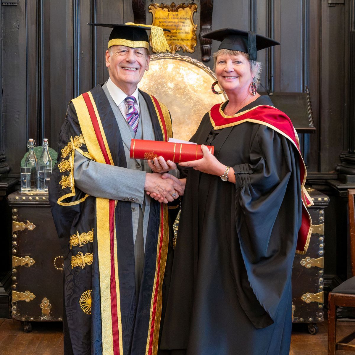 Joy Kettyle dressed in gown and cap receives her CIFE membership in a cylinder shaped container from Lord Lingfield in the Apothecaries Hall