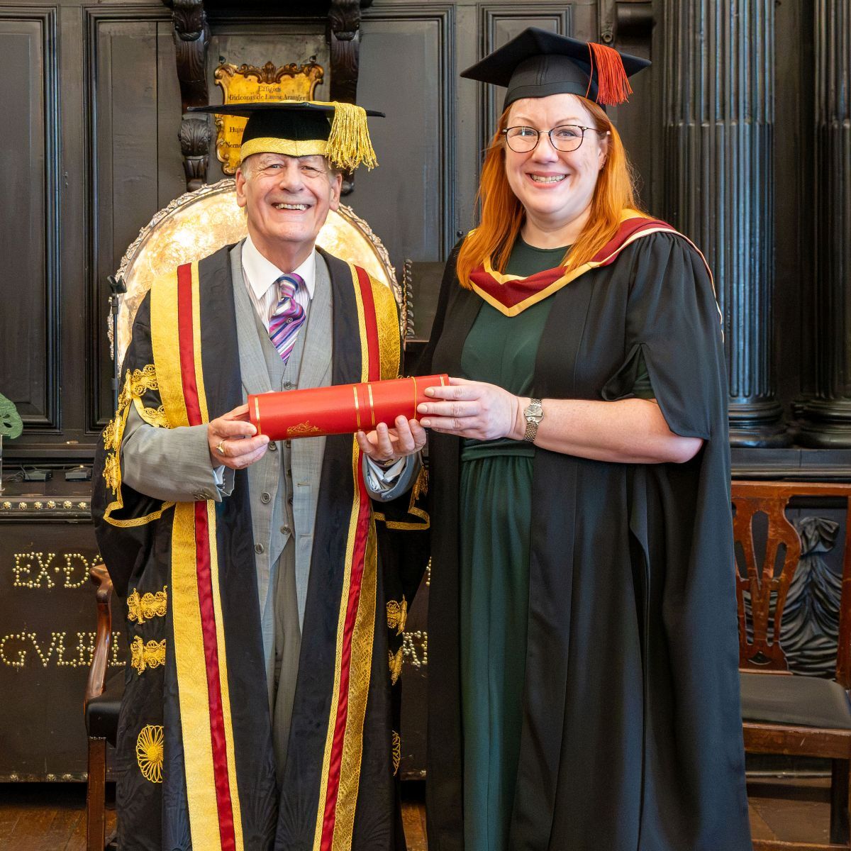 Katharine Dent Jones dressed in gown and cap receives her licentiateship in a cylinder shaped container from Lord Lingfield in the Apothecaries Hall