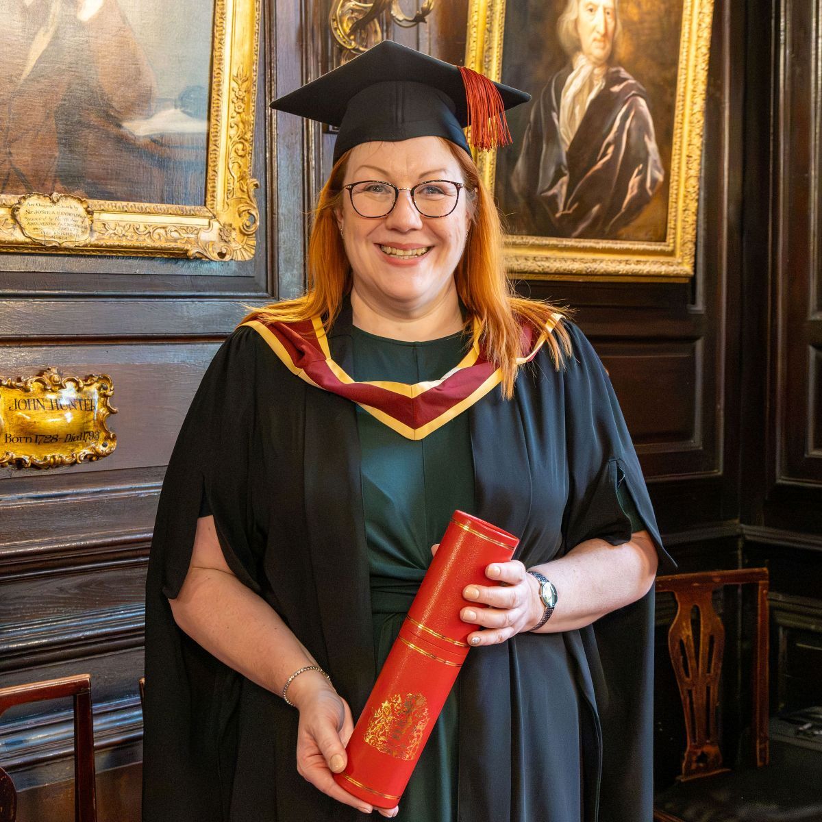 Katharine Dent Jones dressed in gown and cap smiling as she holds her licentiateship in a cylinder shaped container in the Apothecaries Hall