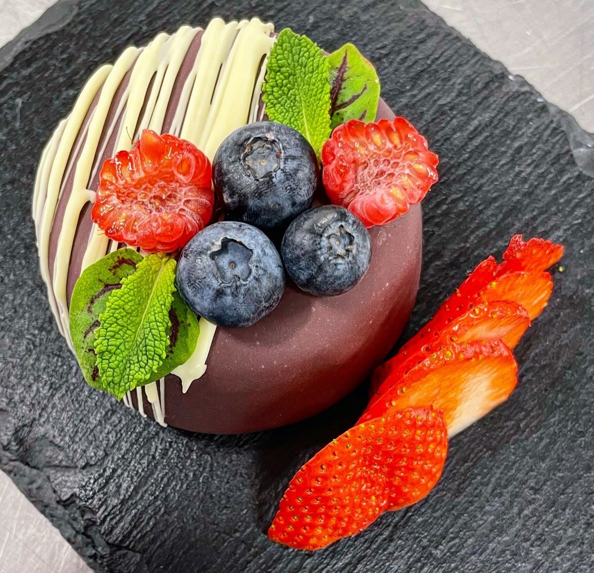 Round dessert covered in milk chocolate with piped white chocolate raspberries blueberries and mint leaves on the top and halved strawberries as a side garnish
