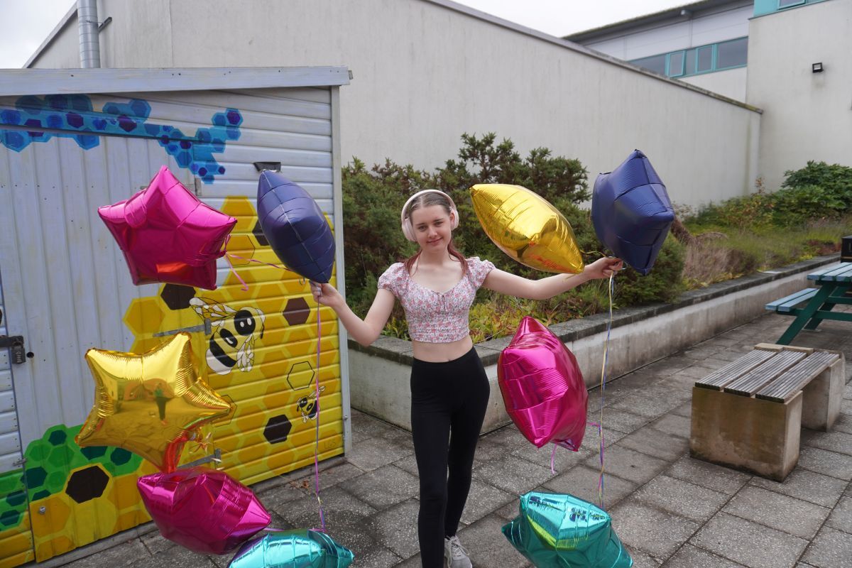 Sian Duffield celebrates her GCSE results holding star shaped balloons which are blowing in the wind in the York College Podium Garden