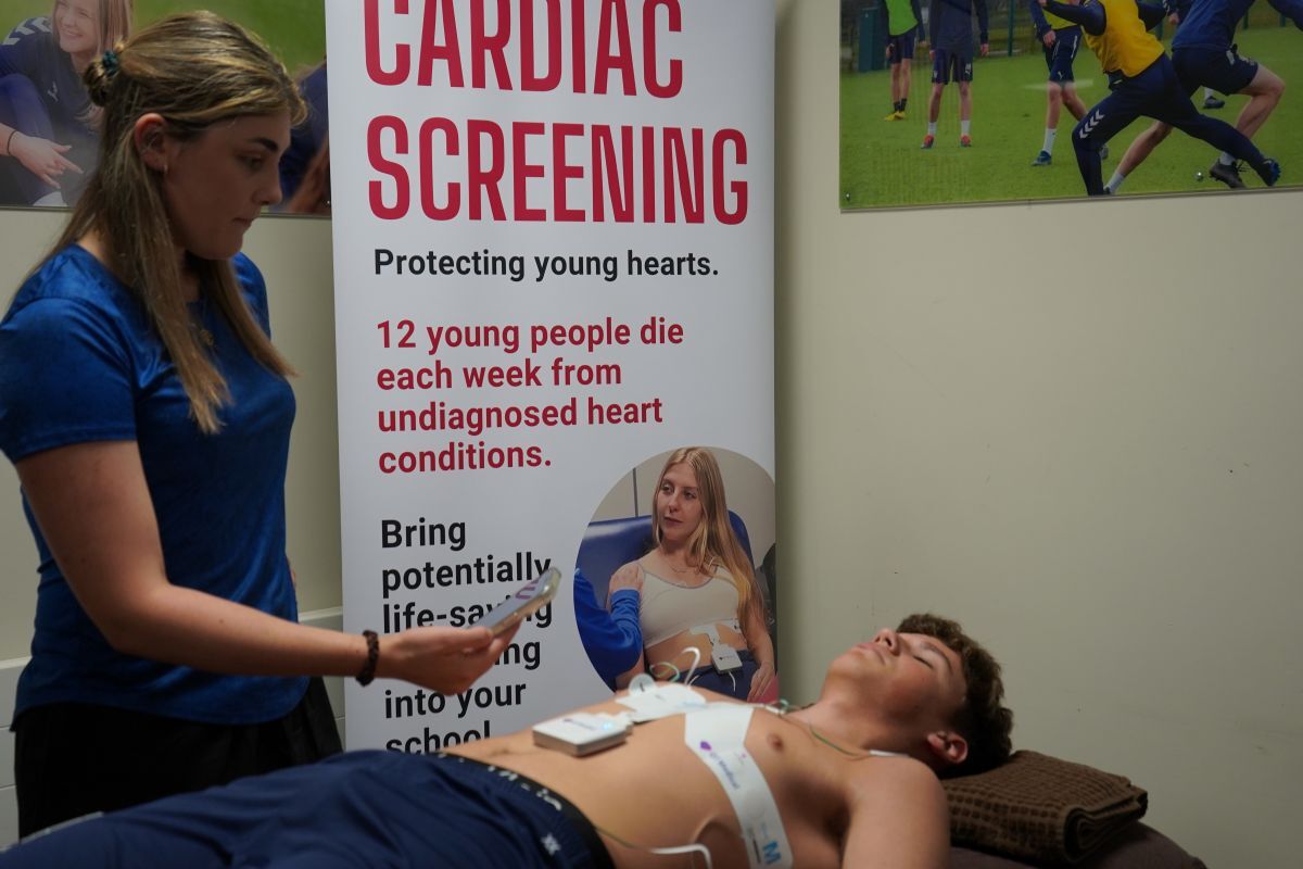 Student lays down on patient bed with ECG electrode patches on as sports tutor Emma Palmer checks readings on mobile device