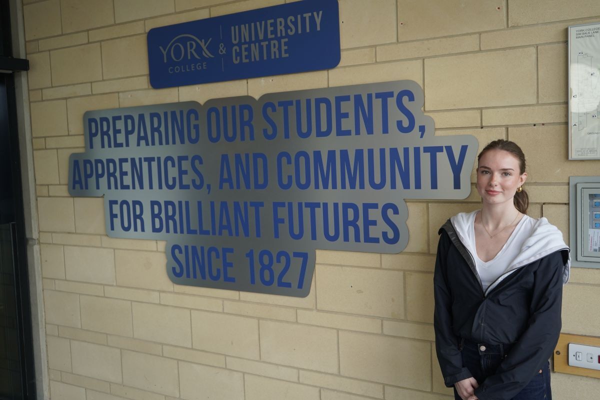 Tilly Allan poses next to Preparing Our Students Apprentices and Community for Brilliant Futures since 1827 wording on brick wall just inside York College entrance