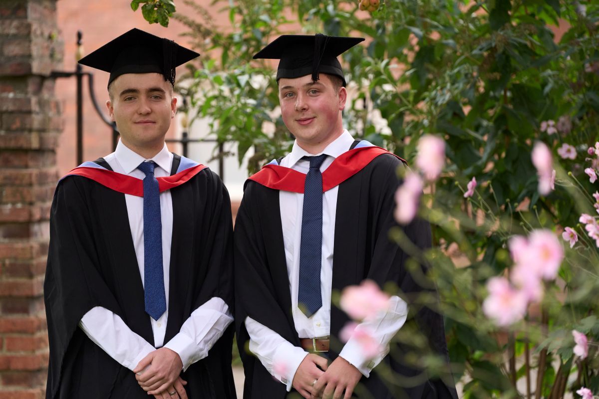 Two male graduates pose in front of iron railings and next to greenery