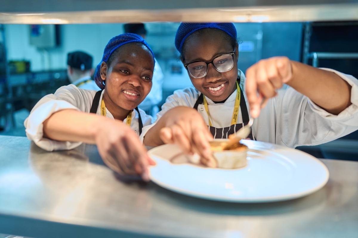 Two professional cookery students are working at the restaurant kitchen serving hatch preparing a plate for service
