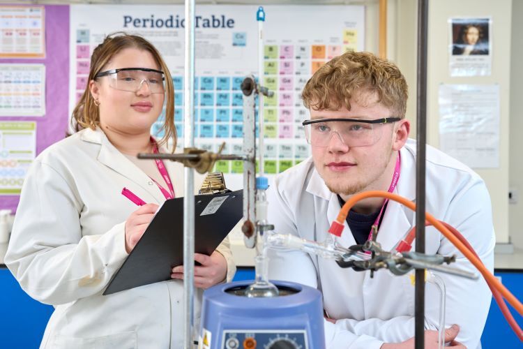 Two students undertake a science experiment in labcoats and goggles