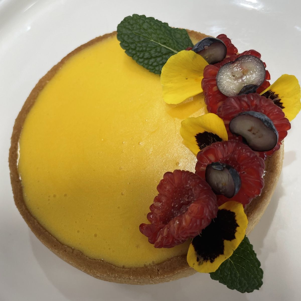 Yellow fruit tart topped with raspberries mint leaves and flower petals down one side