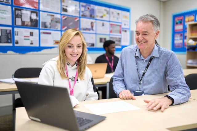 A student and a tutor chat as the student works on a laptop Business