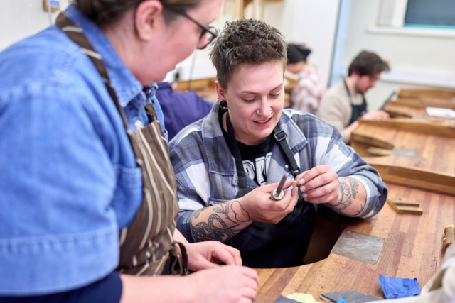 A student uses a chisel to create some jewellery while a tutor looks on