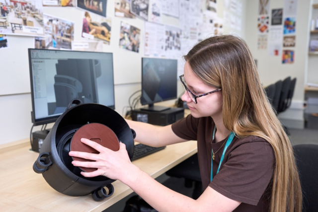 An engineering student holds a product created from their CAD drawing which is on the screen behind them