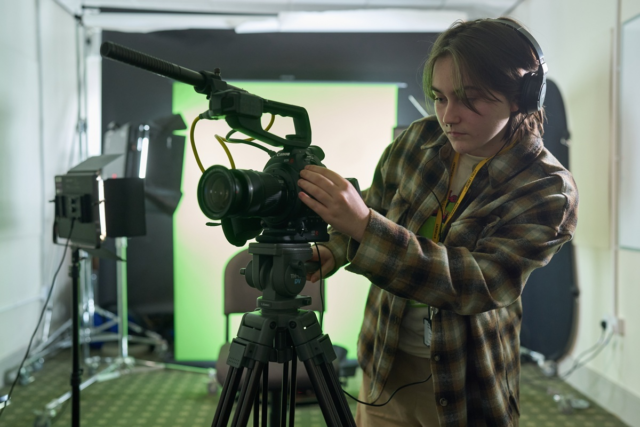 Creative media student wears headphones and is looking into the viewfinder of a camera which is on a tripod