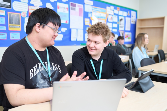 Two Business students sit at a desk looking at each other talking and smiling They have a laptop and tablet on the desk in front of them