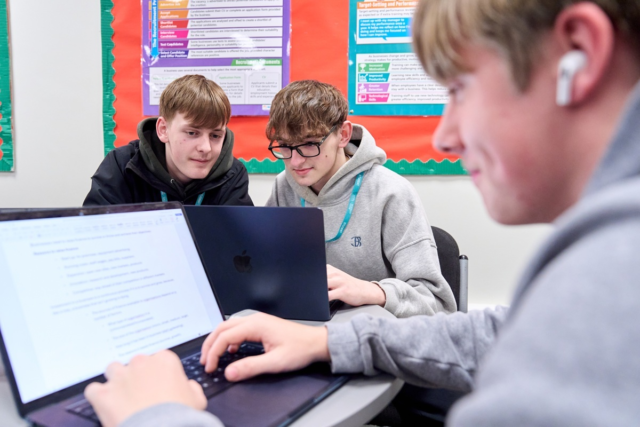 A student working at a laptop with two students sitting behind him discussing work on a laptop