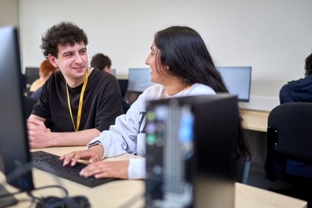 Two students looking at each other smiling as they sit in front of computers