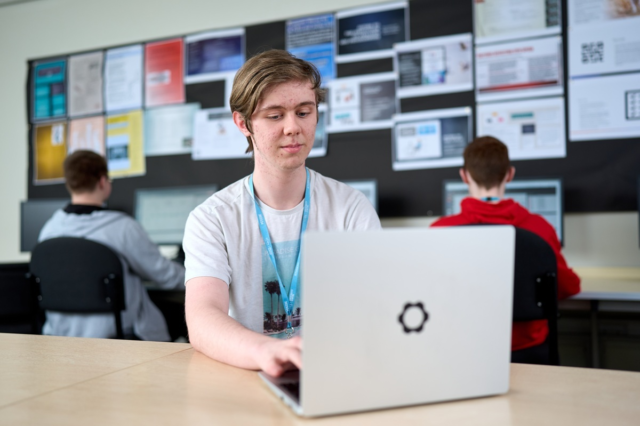 A T Level Digital Sofware Development student sits at desk in front of a laptop with other students working at computers behind them