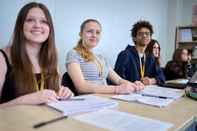 A Level students sitting at a row of desks smiling and looking towards the front of class