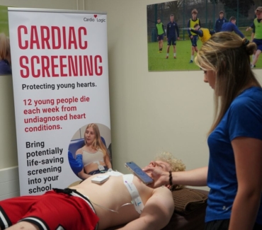 Sports tutor Emma Palmer looks at readings on her mobile phone device as student lies on the patient bed with ECG electrodes attached to his chest