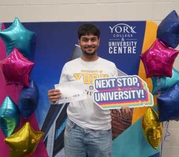 Azaan Iyer celebrates his A Level grades holding his results and a Next Stop University sign in front of banner with the York College building on it and surrounded by star shaped balloons
