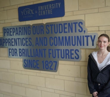Tilly Allan poses next to Preparing Our Students Apprentices and Community for Brilliant Futures since 1827 wording on brick wall just inside York College entrance