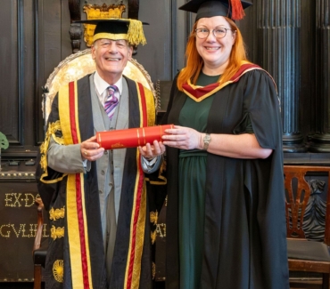Katharine Dent Jones dressed in gown and cap receives her licentiateship in a cylinder shaped container from Lord Lingfield in the Apothecaries Hall